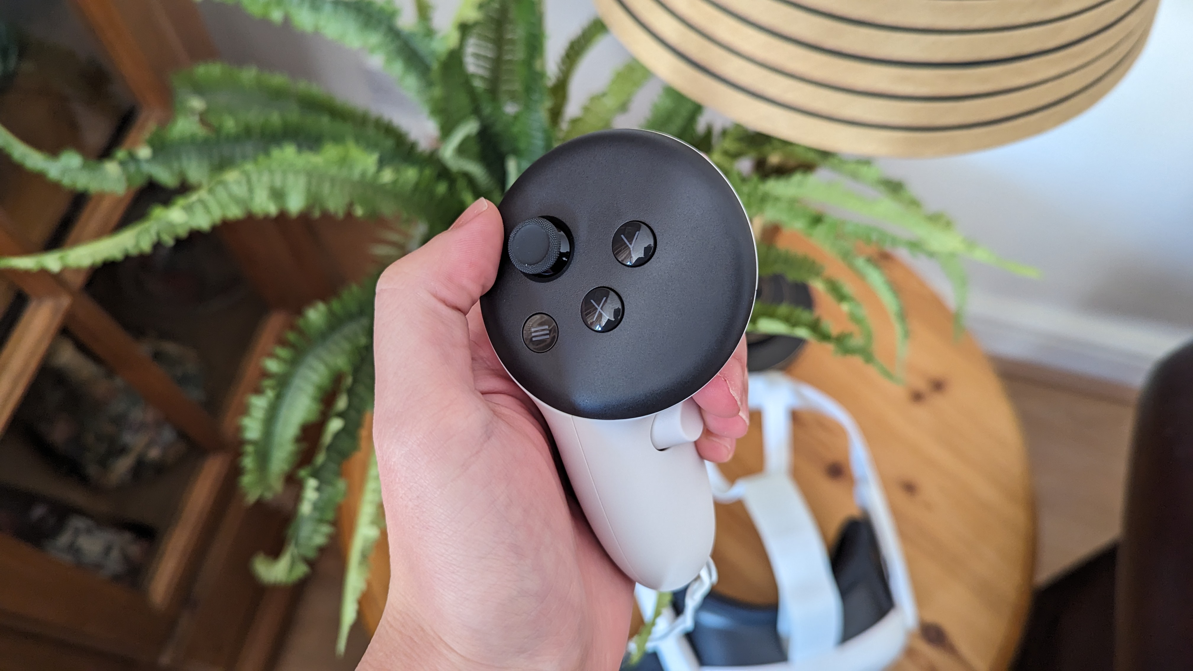 The Meta Quest 3 controller being held above a table with a lamp, a plant and the Quest 3 headset on. You can see the buttons and the thumbstick on top.