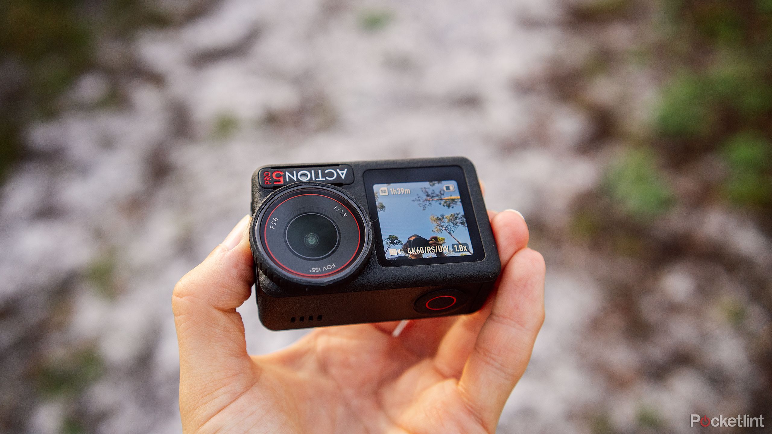 A hand holds the DJI Osmo Action 5 Pro over a sandy trail. 