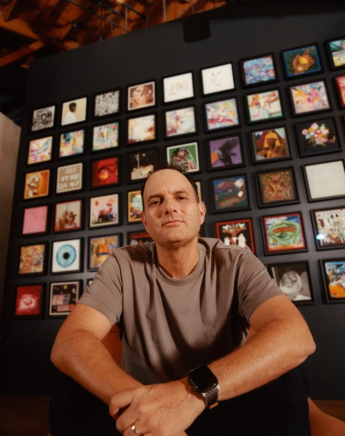 John Janick sitting under a wall display of album covers at Interscope’s offices in LA. He is wearing a grey T-shirt and smiling at the camera