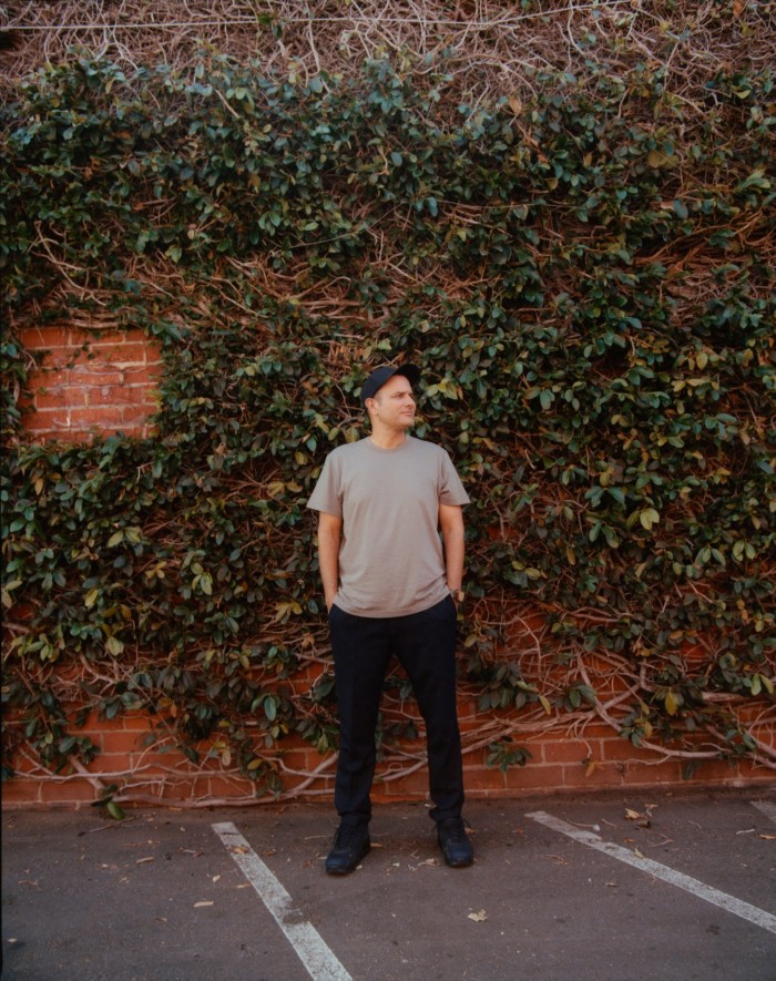 John Janick, the CEO of Interscope, standing by an ivy-clad wall at Interscope’s offices in LA. He wears a grey T-shirt, black trousers and a black baseball cap