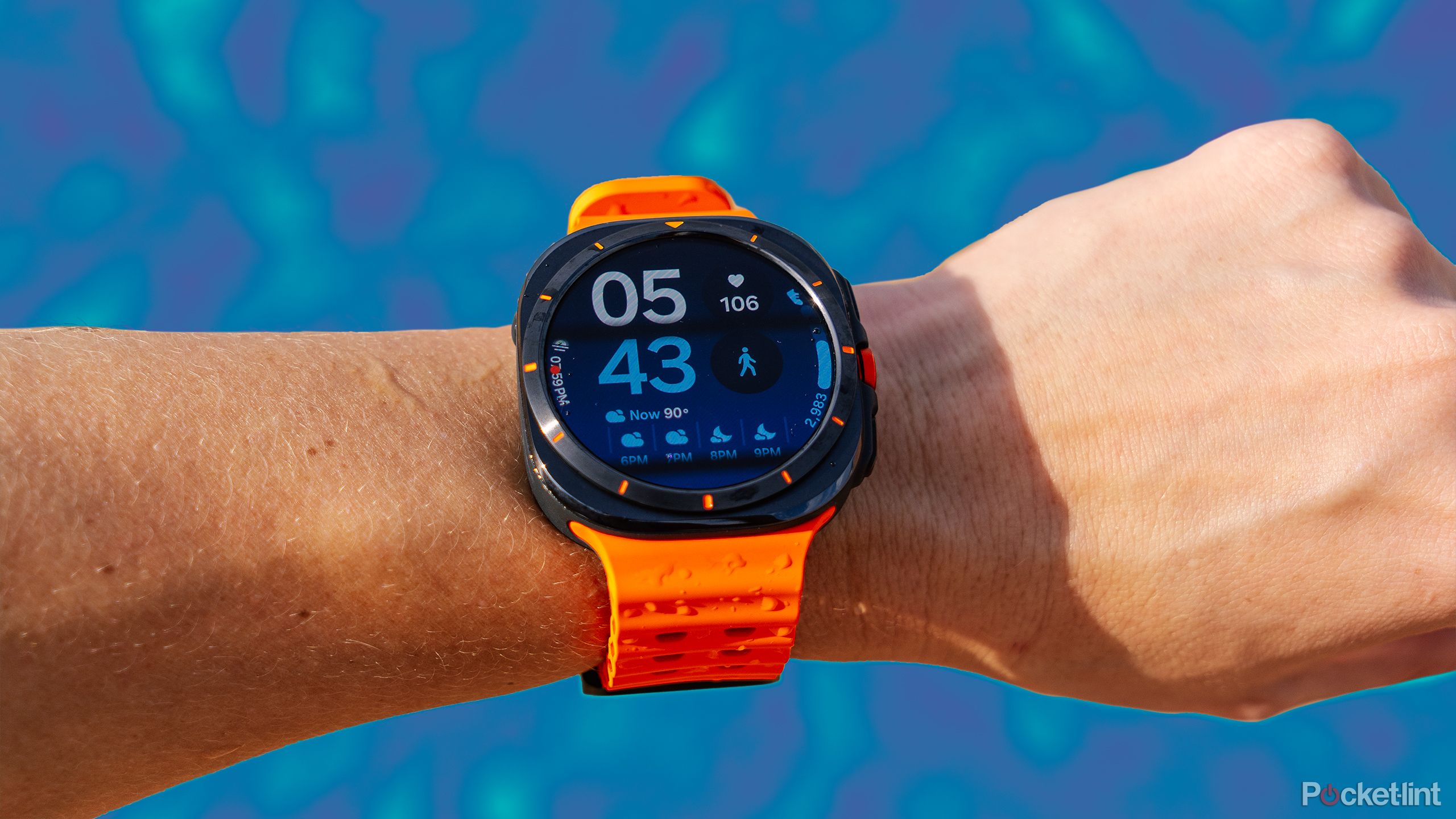 The Samsung Galaxy Watch Ultra is on a wrist held in front of a blurred blue background. 