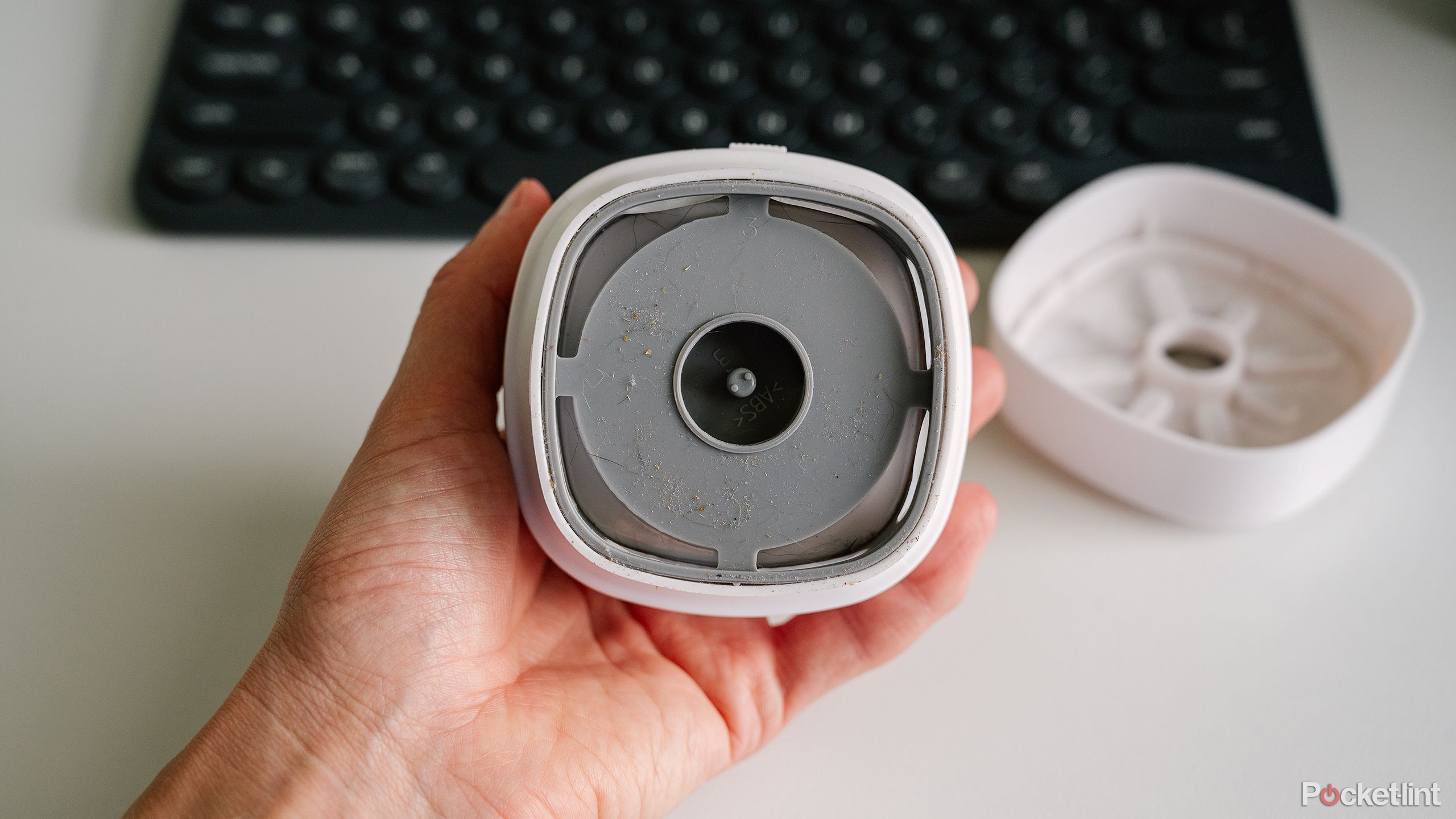 A hand holds the Odistar Desktop Vacuum cleaner with the dust bin open over a white desk and keyboard.