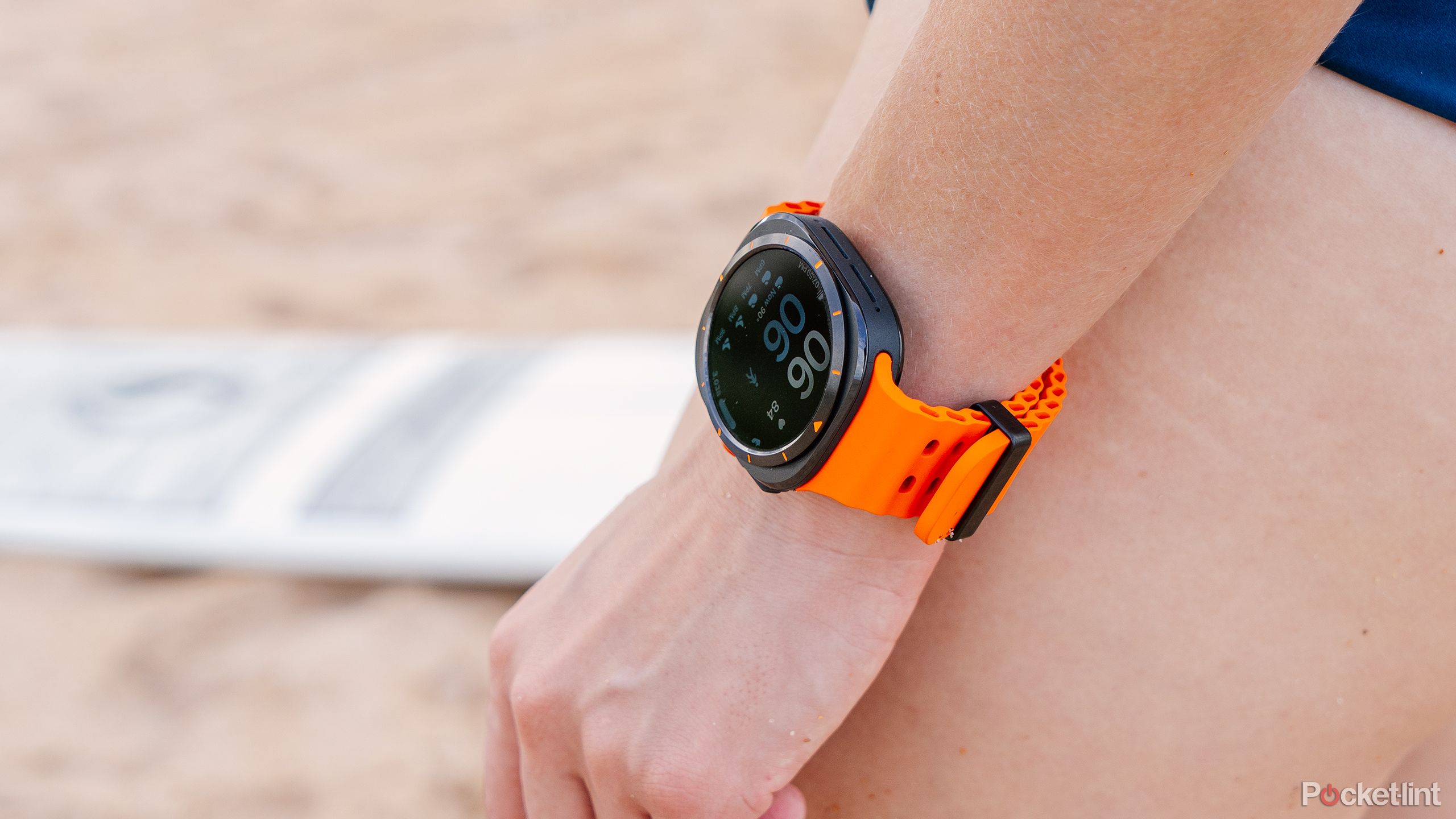 The Samsung Galaxy Watch Ultra on a wrist that rests on a leg while they stand on a beach. 