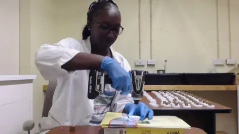 Shamika Spencer, wearing glasses, a white lab coat and blue gloves, is holding a drill in a lab where she conducts experiments. In the background, a large table holds what appear to be samples.