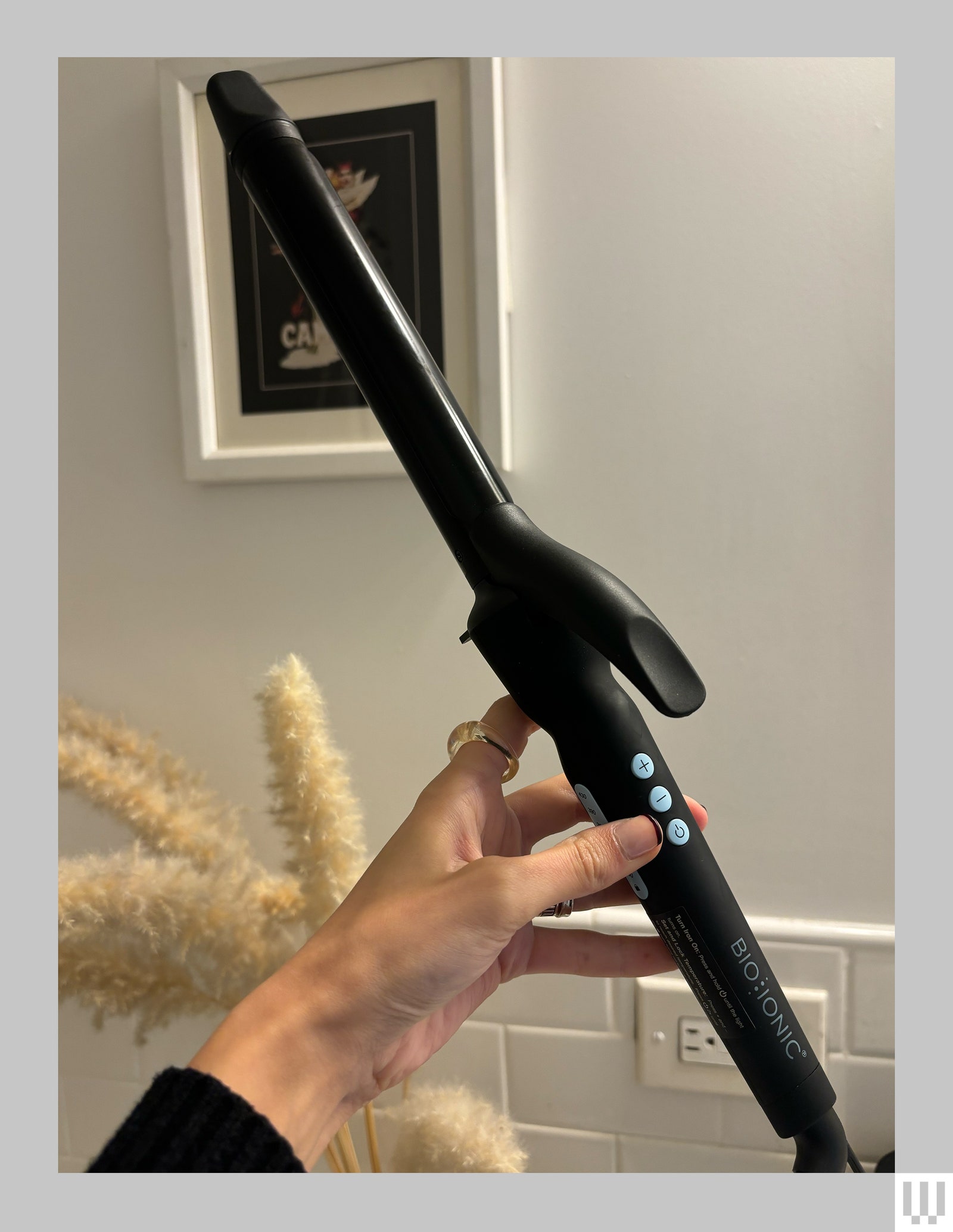 Side view of a hand holding the Bio Ionic Long Barrel Styler a long narrow black curling iron with three 3 small light...