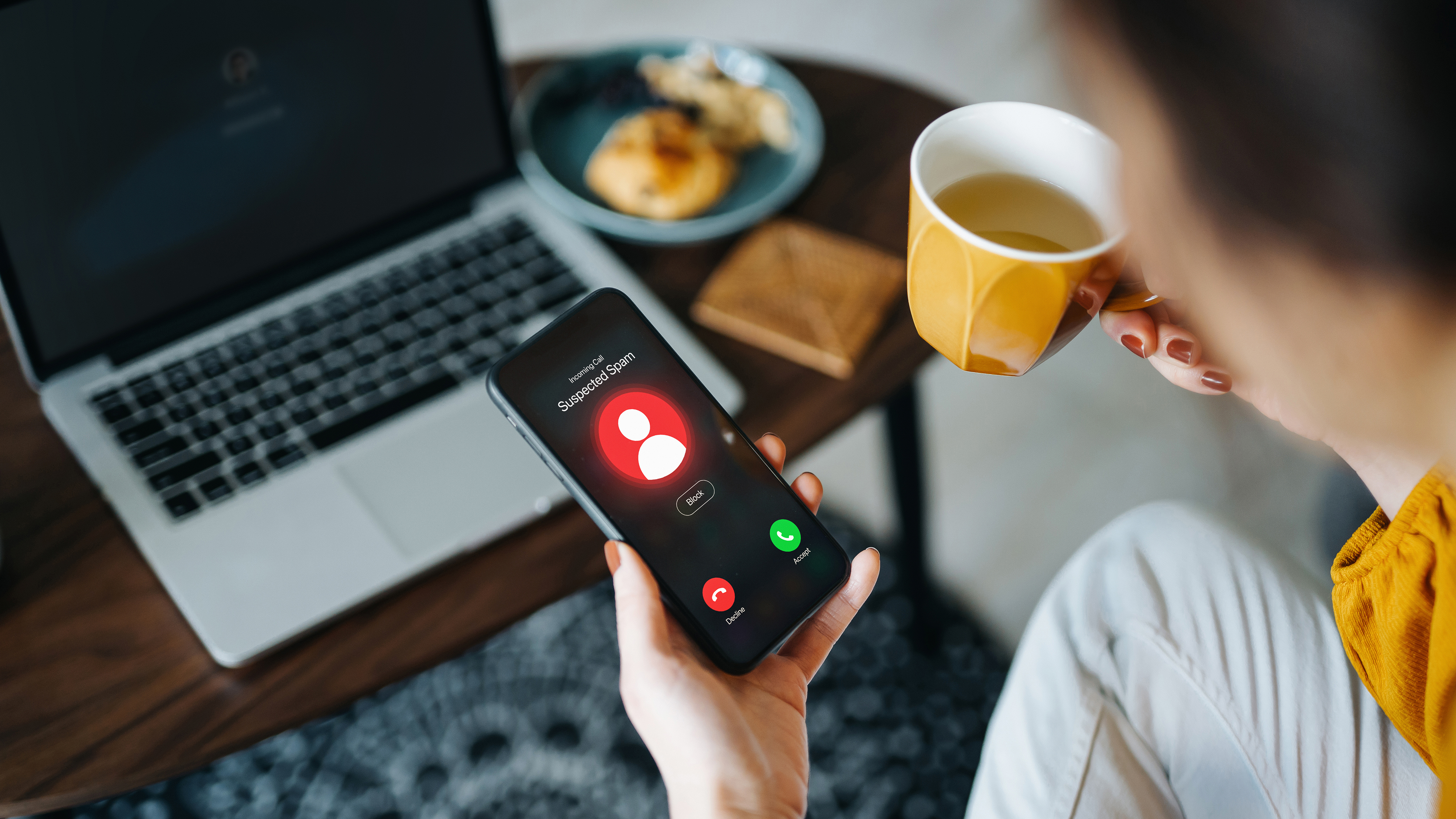 A hand holding a phone showing a suspected scam phone call, next to a hand holding a mug