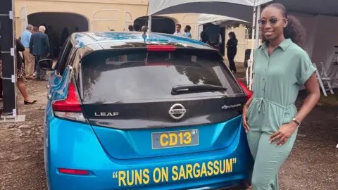 BBC Biologist Shanika Spencer poses next to a blue Nissan Leaf car on which the sentence "Runs on Sargassum" is printed. 