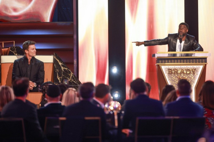 A man in a black suit is seated on stage, smiling, while another man in a leather jacket gestures and speaks at a podium labeled ‘Brady.’ The audience is seated in the foreground