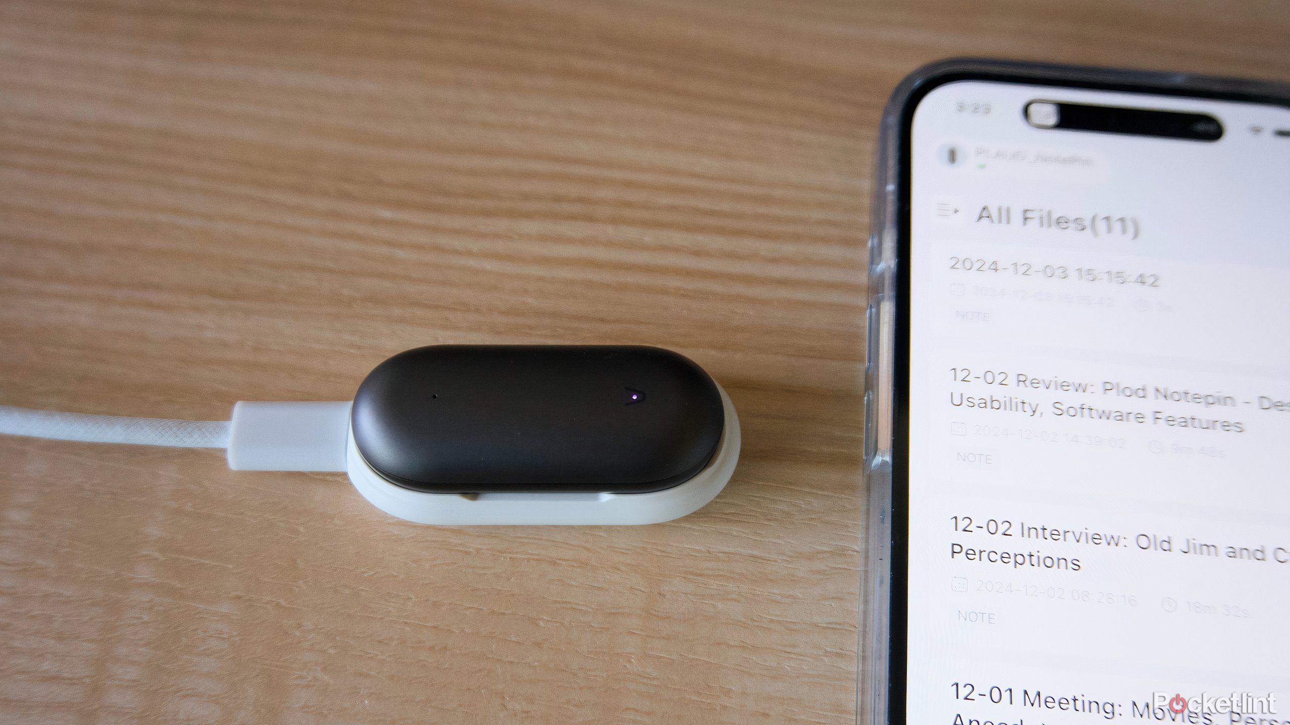 A Plaud NotePin charging on a white charging dock next to an iPhone 16.