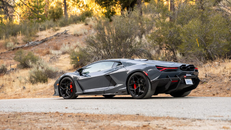 A sleek, gray sports car with black rims and red brake calipers is parked on a rural road surrounded by dry shrubs and trees.