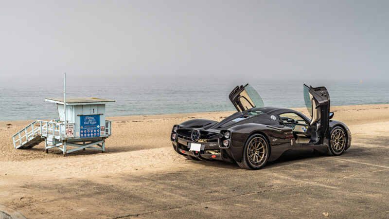 A sleek black exotic car with gull-wing doors open is parked near a beach, with a lifeguard station quietly standing in the background.