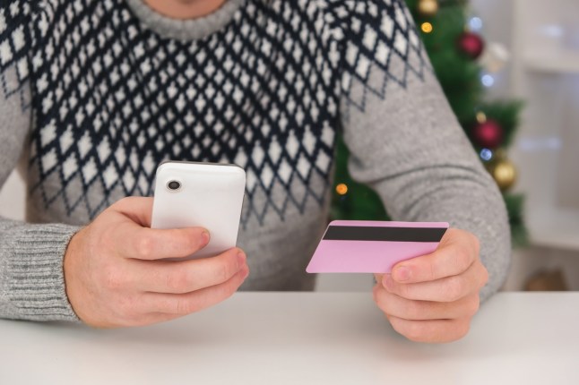 A man makes online purchases using his phone the New Year holiday. A man orders Christmas gifts using his phone for Christmas and pays them with a credit card. The man holds credit card in his hand.