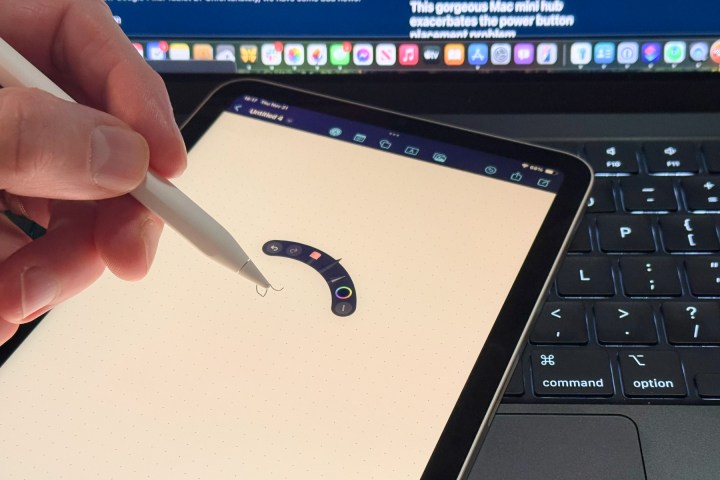 An Apple Pencil Pro being held in a person's left hand with squeeze controls showing on an iPad mini 7 on top of a MacBook Pro keyboard.