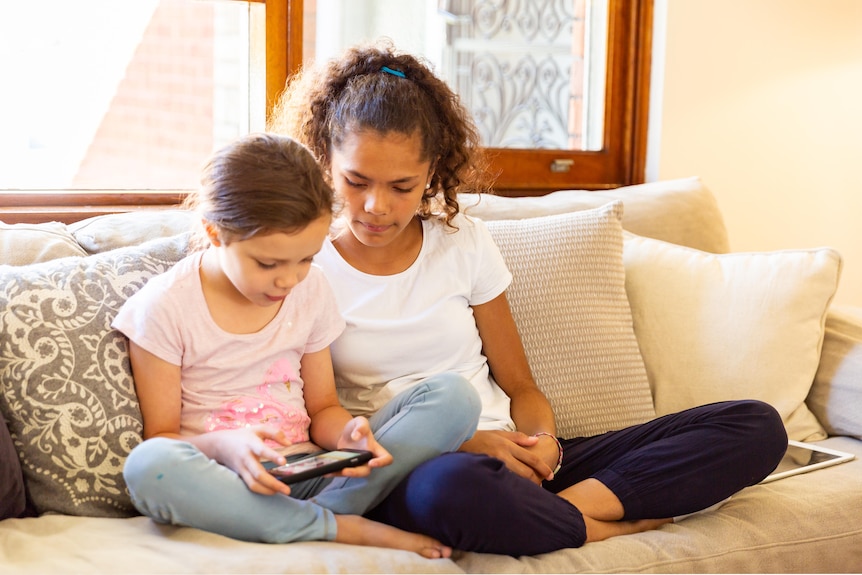 Two sisters aged under 13 are sitting on a sofa looking at a smart phone. They are both interested in what is on the phone