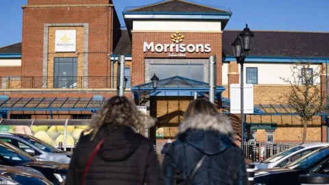 Getty Images Two women in coats walking into a large Morrisons store with the logo above the entrance