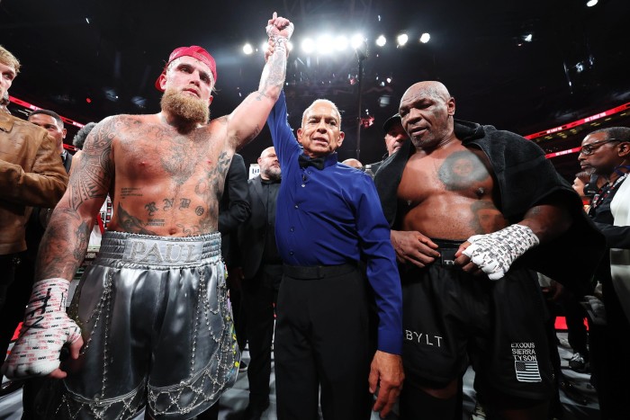 A victorious boxer with tattoos and silver shorts labeled ‘Paul’ has his arm raised by a referee in a boxing ring. Another boxer with a towel draped over his shoulders stands beside them, showing a tattoo on his chest