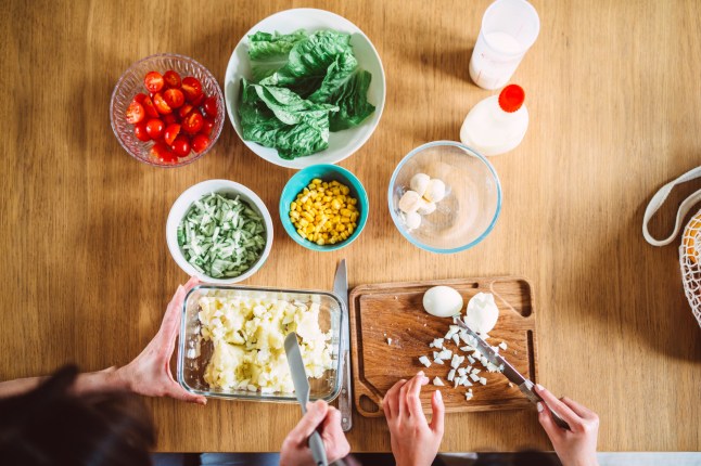 Directly above shot of a mother and daughter in the process of preparing a fresh and healthy salad. The image shows a variety of colourful vegetables and ingredients neatly arranged on a wooden table, including lettuce, cherry tomatoes, cucumbers, boiled eggs, and sweetcorn. Hands are seen slicing cucumbers on a cutting board, emphasising teamwork and the joy of cooking together. Concepts of family bonding, healthy eating, and culinary creativity.