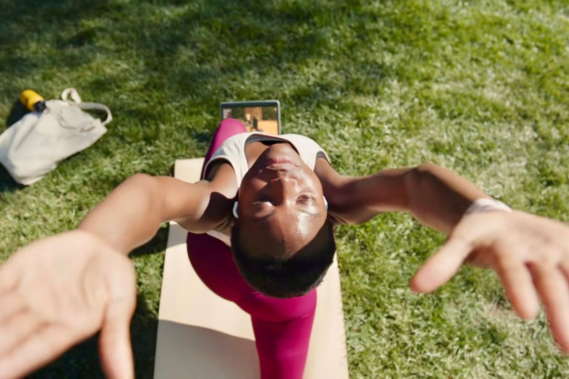 An individual using Apple Fitness+ to do yoga in the park on the grass with a yoga mat and an iPad.