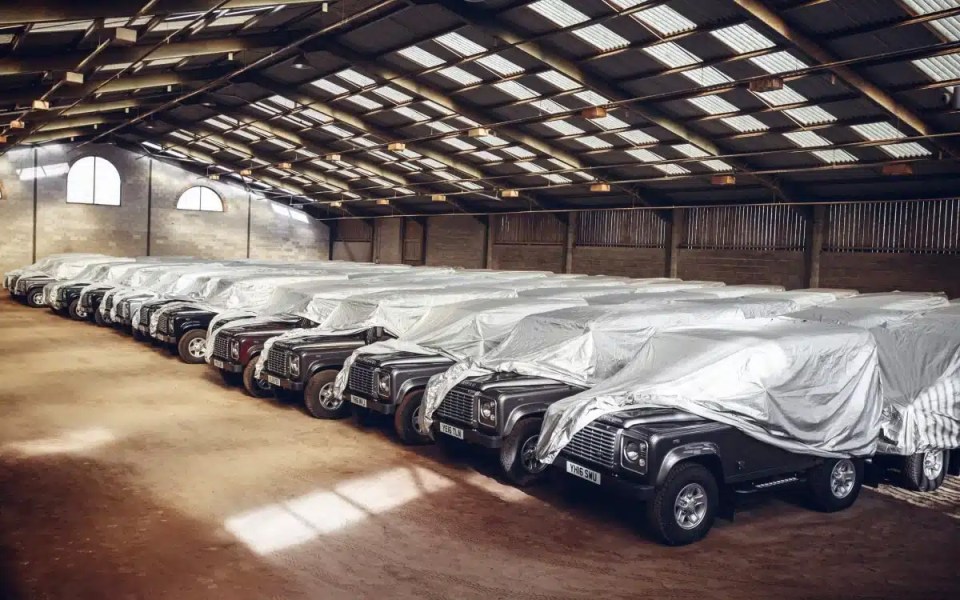 Many Land Rover Defenders covered in protective sheets inside a large warehouse.