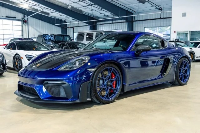 A stunning blue Porsche 718 Cayman GT4 RS with black accents is parked in a showroom, catching every eye. Other cars are visible in the background, highlighting this exclusive Porsche for sale.