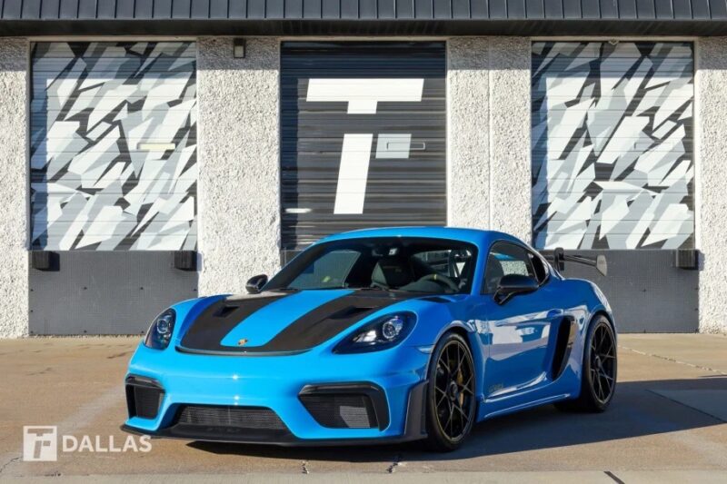 A blue Porsche 718 Cayman GT4 RS with black accents is parked in front of garage doors featuring geometric patterns, and it's now for sale.