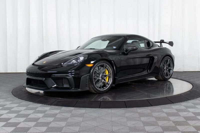 A sleek black Porsche 718 Cayman GT4 RS, complete with a spoiler, is parked on a checkered floor against a white backdrop. Perfect for enthusiasts looking to buy performance and style in one package.