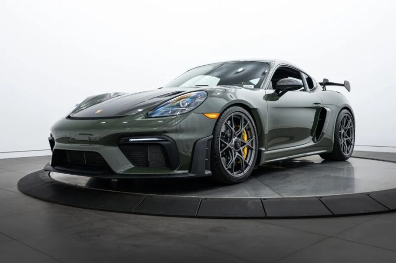 For sale: A stunning dark green Porsche 718 Cayman GT4 RS on a rotating platform, showcasing a sleek design, aerodynamic body, large rear spoiler, and black alloy wheels. Don't miss the chance to buy this exceptional sports car.