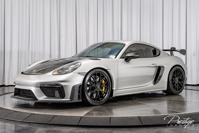 The Porsche 718 Cayman GT4 RS, a silver sports car with black detailing and yellow brake calipers, is showcased elegantly in a showroom with a white curtain backdrop. This masterpiece is for sale to discerning buyers seeking unmatched performance and style.