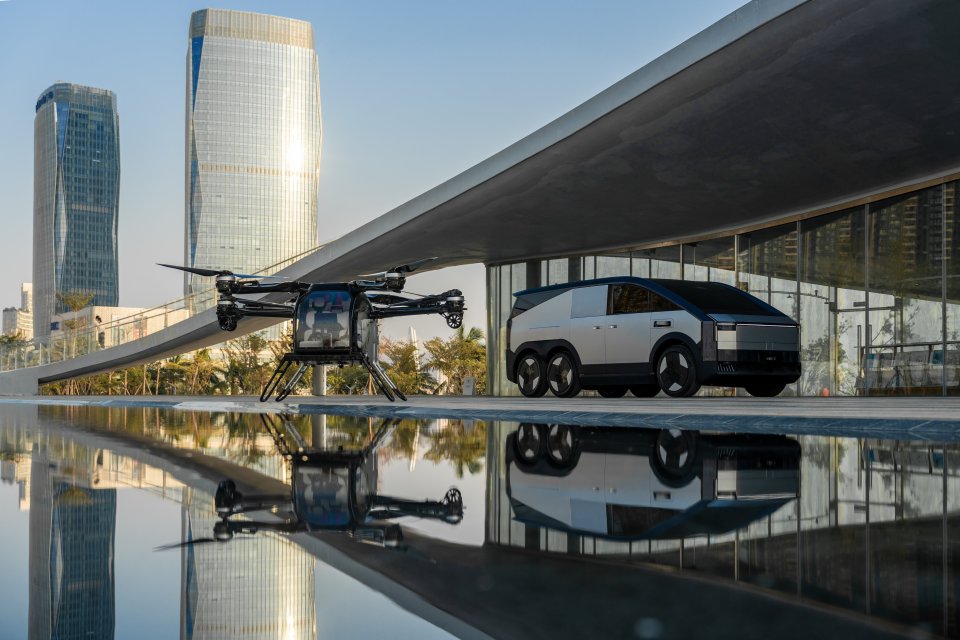 Six-wheeled vehicle and a flying drone parked near a reflecting pool.