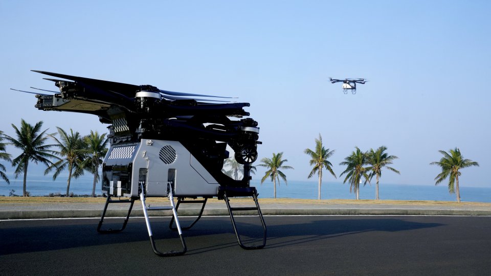 Six-wheeled truck with a built-in flying machine and a smaller drone in flight.