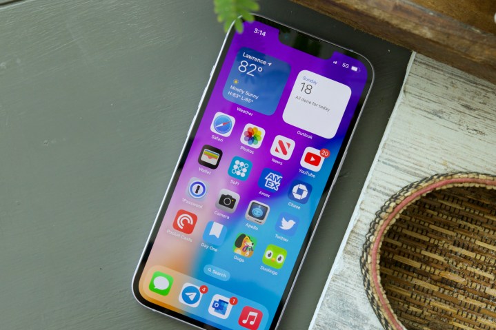 An iPhone 14 laying on a table. The display is on and showing the home screen.