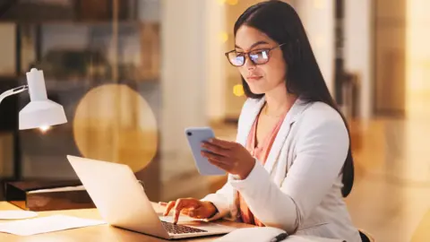 Getty Images A woman sitting at her computer and using her phone