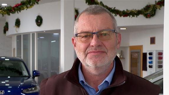 Miles Jude standing in a car sales room. He has short grey hair and glasses. he is wearing a vlue shirt and a brown jacket. 