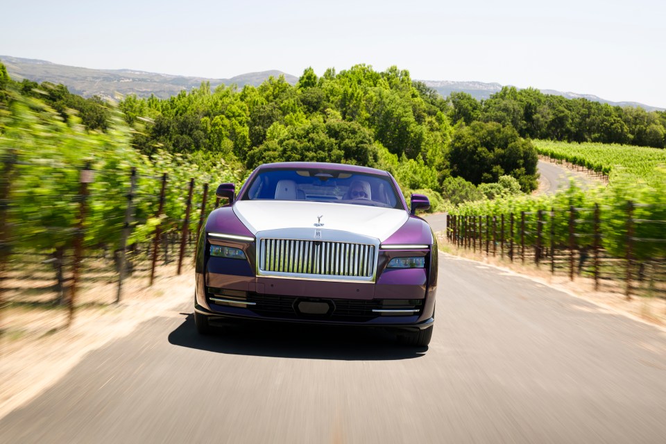 A purple Rolls-Royce Spectre driving on a road through vineyards.