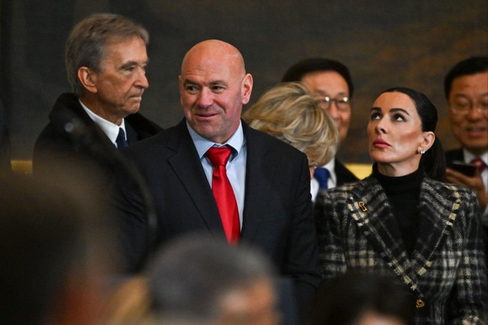 A smiling confident-looking shaven-headed man wearing a red tie makes his way through the crowd to his seat