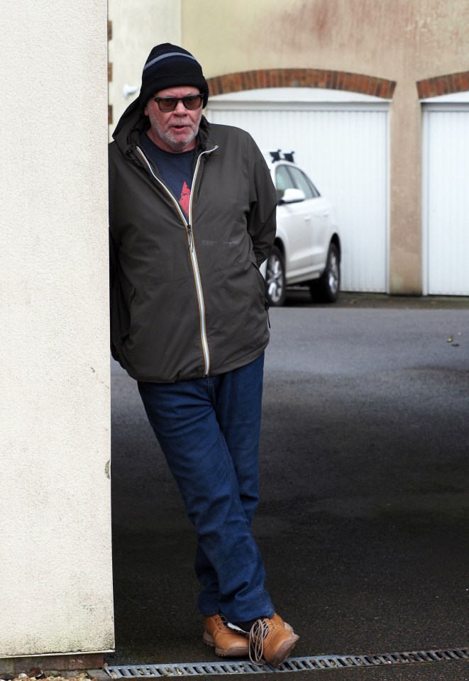 A man standing near a wall, looking towards the camera.  A car is visible in the background.