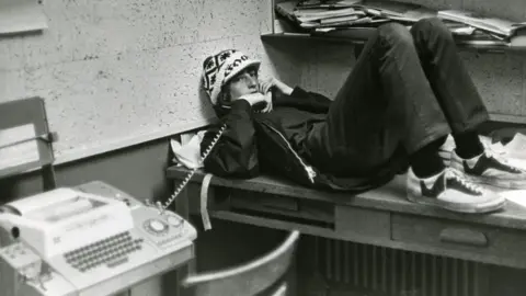 Lakeside School A teenage Bill Gates wearing a hat, lying down on desk, leaning against wall as he holds a landline phone, in a black and white photograph