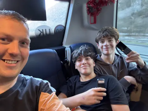 The Fulham Boys School David Smith takes a selfie with two students, who are holding their brick phones and smiling, at the back of a school coach. 