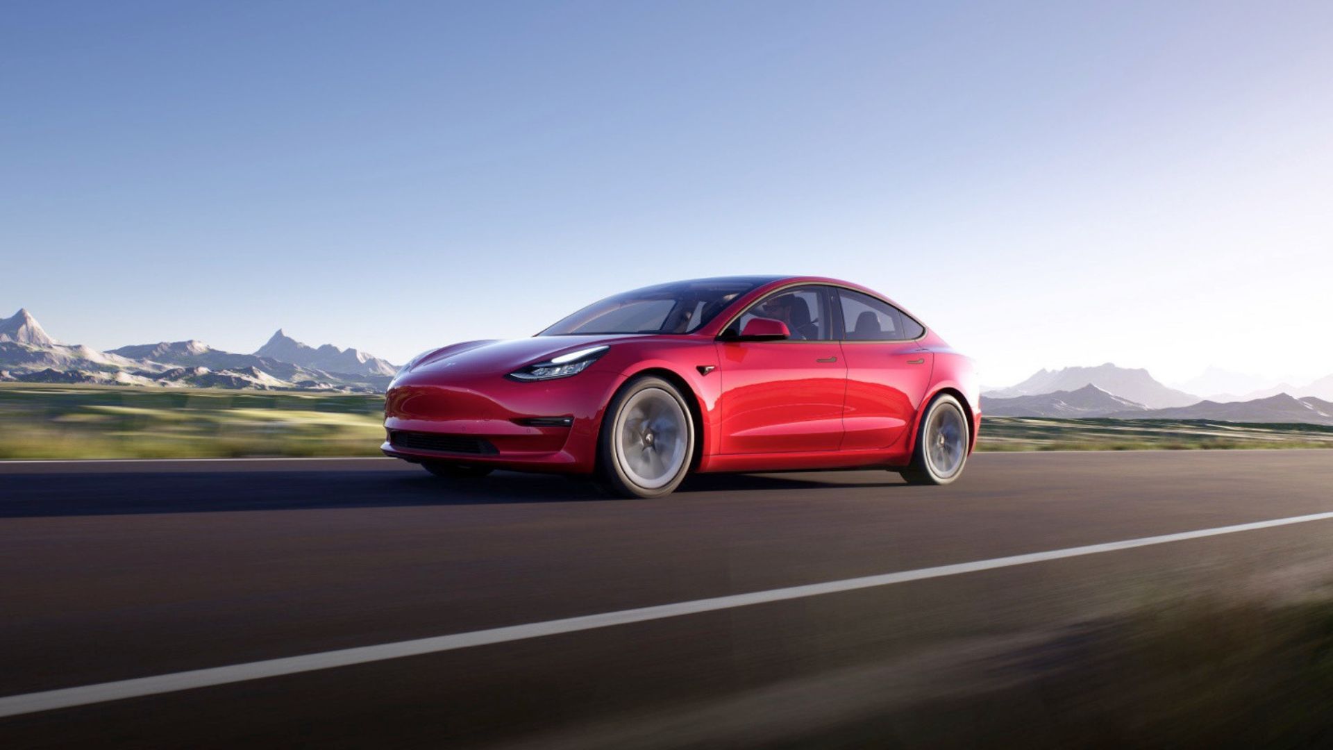 A red Tesla Model 3 driving on a country road with snowcapped mountains in the background.