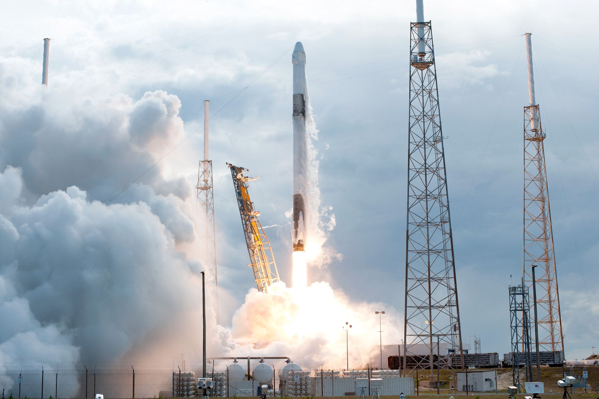 A rocket launches against a cloudy sky, surrounded by tall launch structures and smoke.