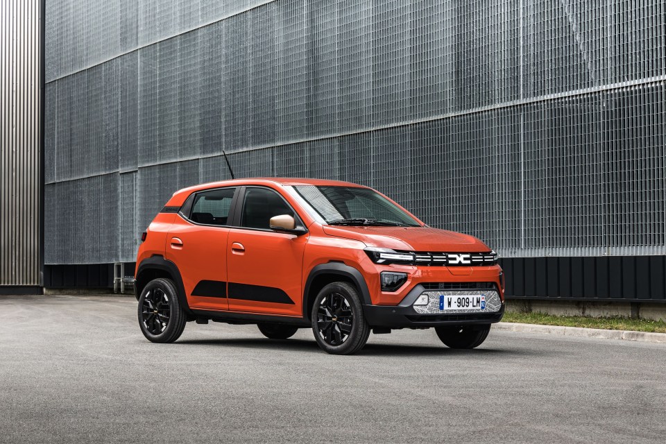 Orange Dacia Spring electric car parked in front of a metal wall.