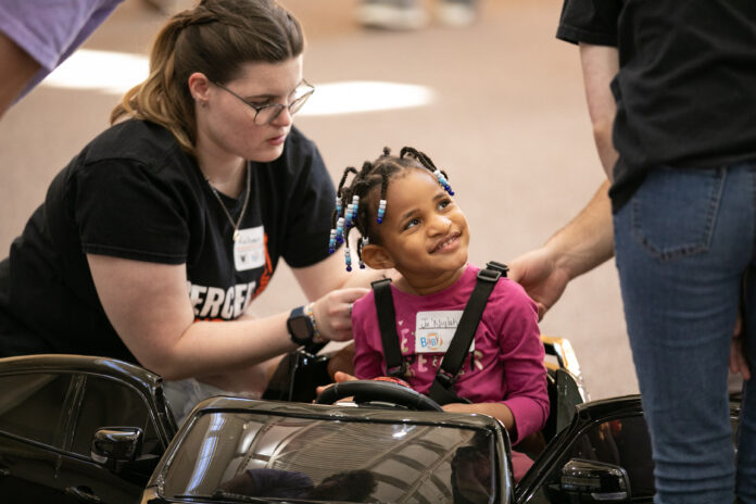 Child in a toy car is helped by two adults. The child is smiling and wearing a pink shirt and name tag, while one adult adjusts the seatbelt.