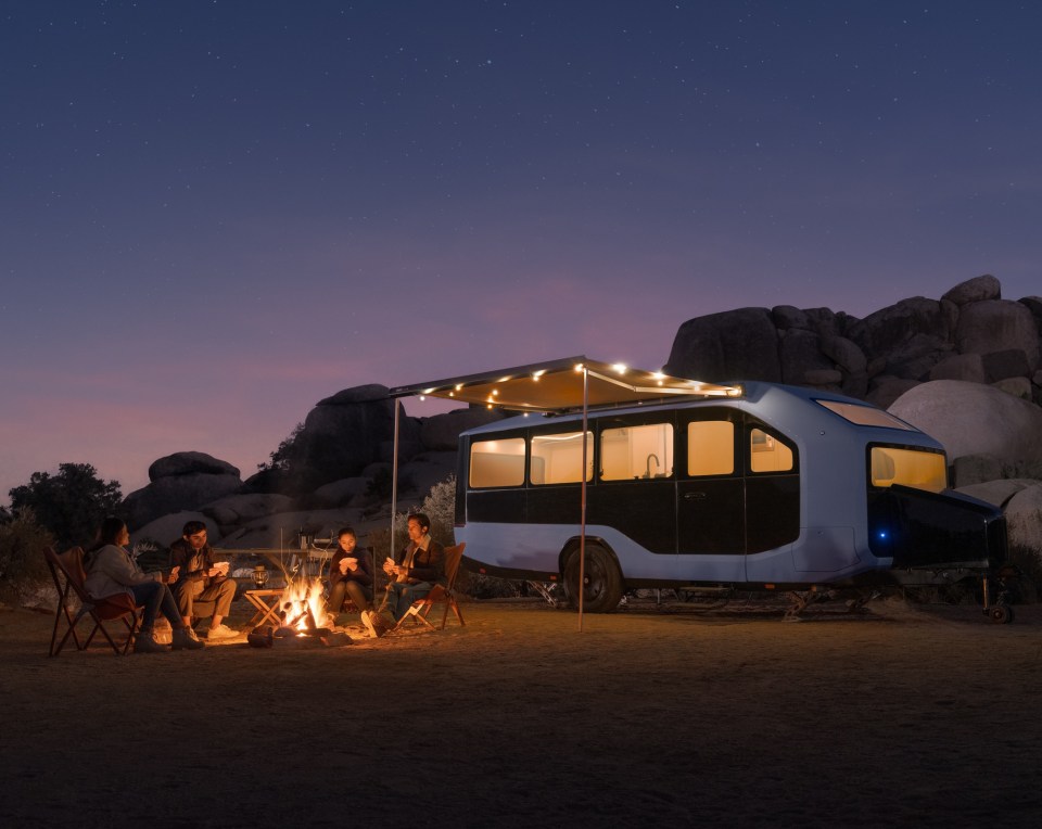 People sitting around a campfire at night next to an electric caravan.