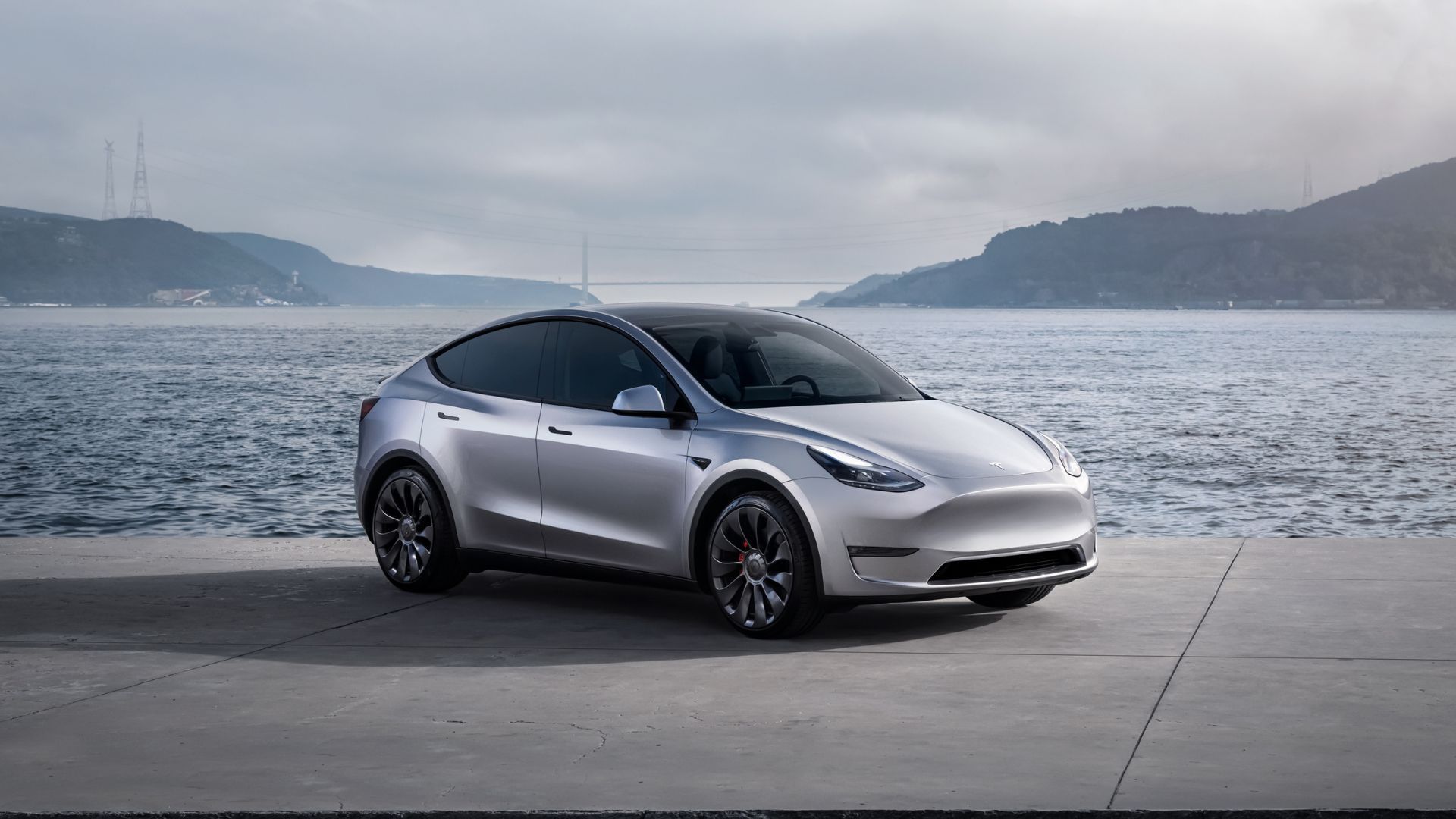 A silver Tesla Model Y parked on tarmac with mountains and bay of water behind it.