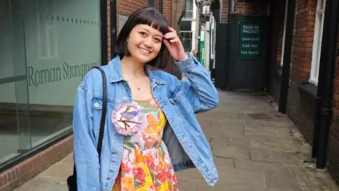 Nina Nina , a young woman smiling at the camera. She is wearing a floral colourful dress with a denim jacket over it.