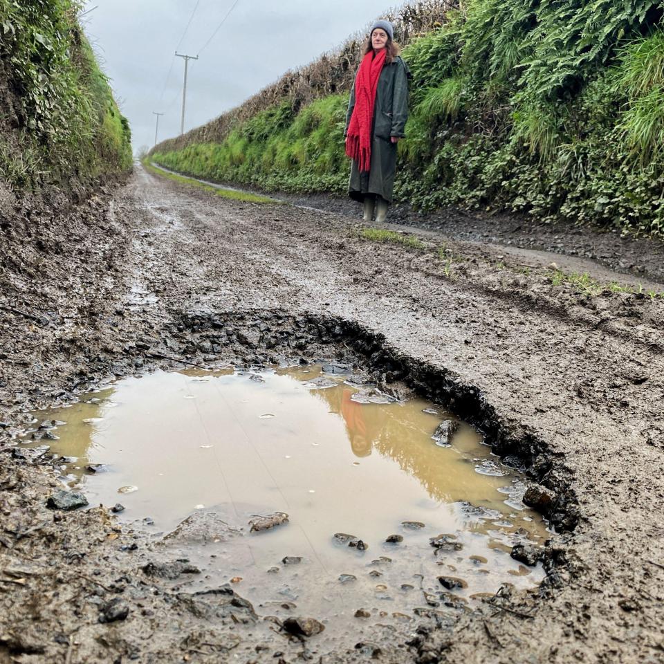 Woman standing by a large pothole in a muddy country lane.