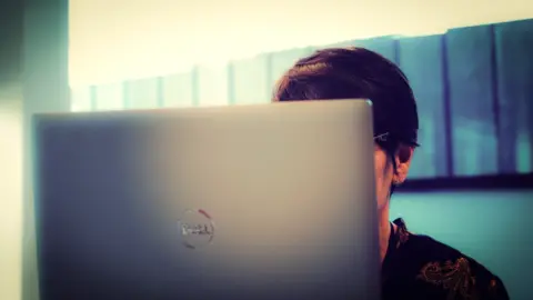 BBC An anonymous lady sits behind a computer laptop screen, you can only see her short dark hair over the top of the laptop