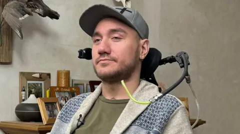 BBC Noland. He is sitting in a wheelchair and has a tube that can be blown into to operate it. He is sharply dressed with a green shirt, white jacket and a grey baseball cap. He has well kept short hair and stubble.