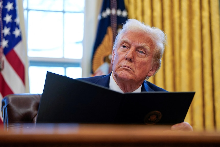 Donald Trump sits at a desk holding a black folder open with yellow-gold curtains and two flags behind him.